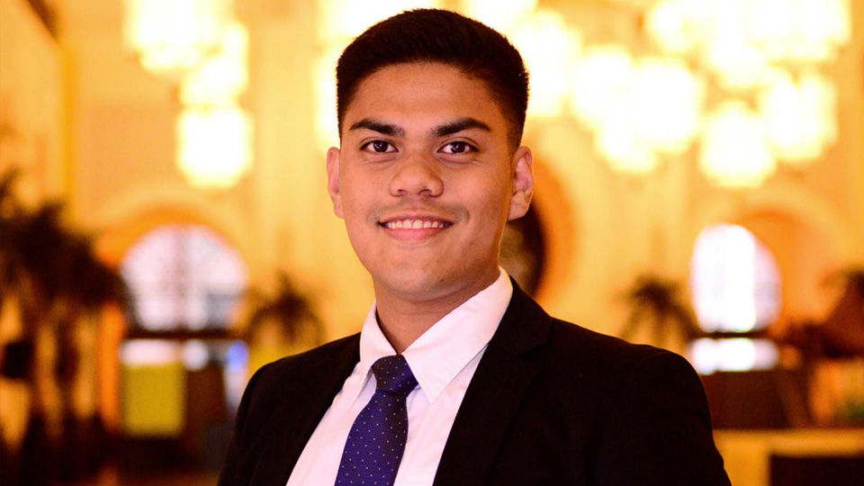 Photo of Angele De Guzman standing in a hotel lobby. He is smiling and is wearing a black suit, white shirt and blue tie