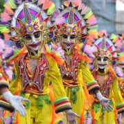 MassKara Festival Article Feature Image. A parade of festival dancers wearing white masks with gold brocade under the eyes and a red painted smiling mouth. A gold crown head piece with red fire tips and yellow square at the center. costumes of yellow material with red, gold, green and black brocade cuffs and chest pieces