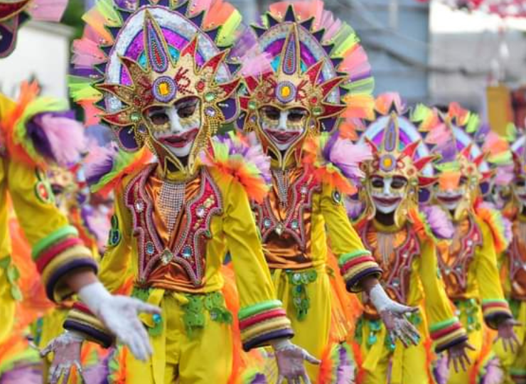 MassKara Festival Article Feature Image. A parade of festival dancers wearing white masks with gold brocade under the eyes and a red painted smiling mouth. A gold crown head piece with red fire tips and yellow square at the center. costumes of yellow material with red, gold, green and black brocade cuffs and chest pieces