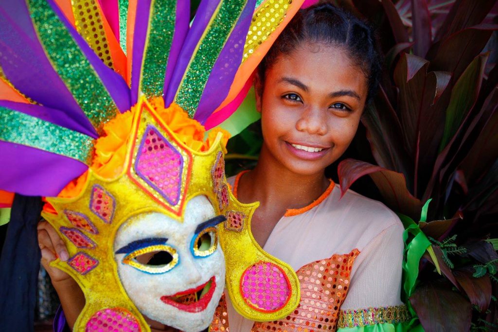 A young filipina (girl) holding a white MassKara festival mask with yellow velvet headdress and vibrant strips of colour fanning out from the forehead of the mask.