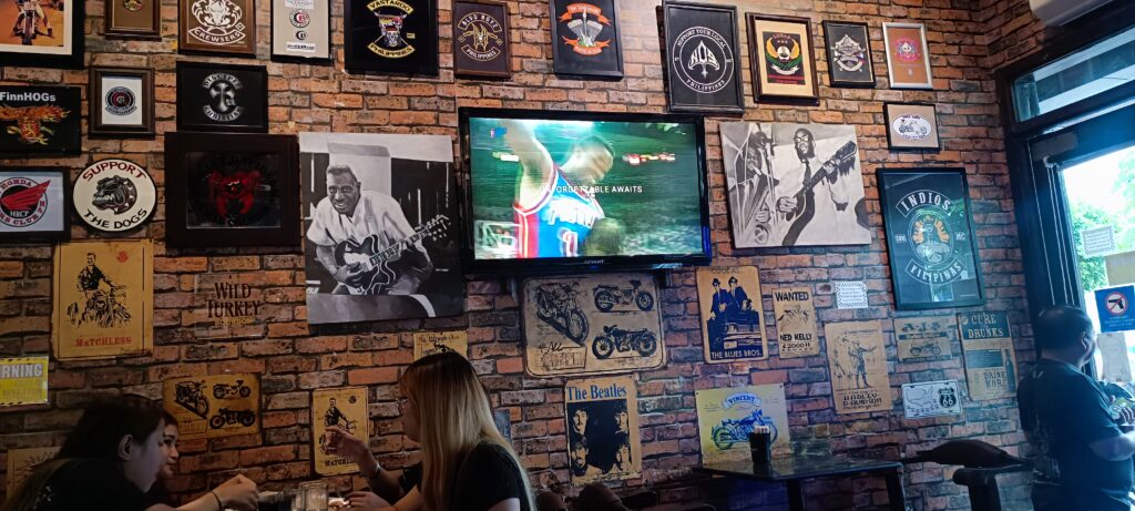 A photo of a brick-effect wall featuring framed photos of Band logos, bands and musicians, with diners seated in front at a table at The Roadhouse Manila Bay