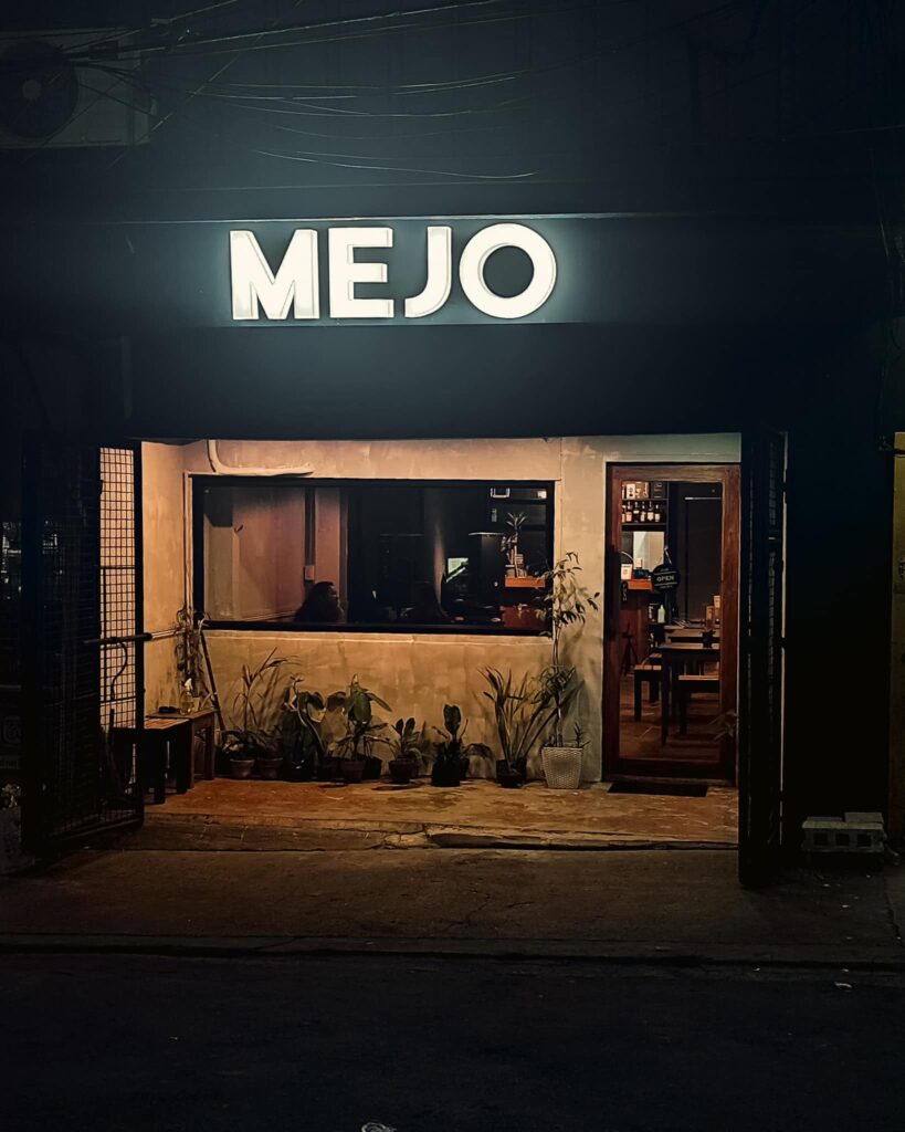 A photo showing the front entrance of MEJO Kitchen & Bar, Quezon City Philippines.  The MEJO signage is lit up and there is a wide horizontal windown to the left of the entrance door.  Plants are below the window and there is a wooden bench against the wall on the left